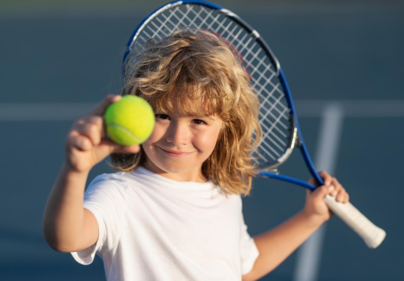 Bambino con racchetta da tennis e pallina, sorridente su un campo da tennis.