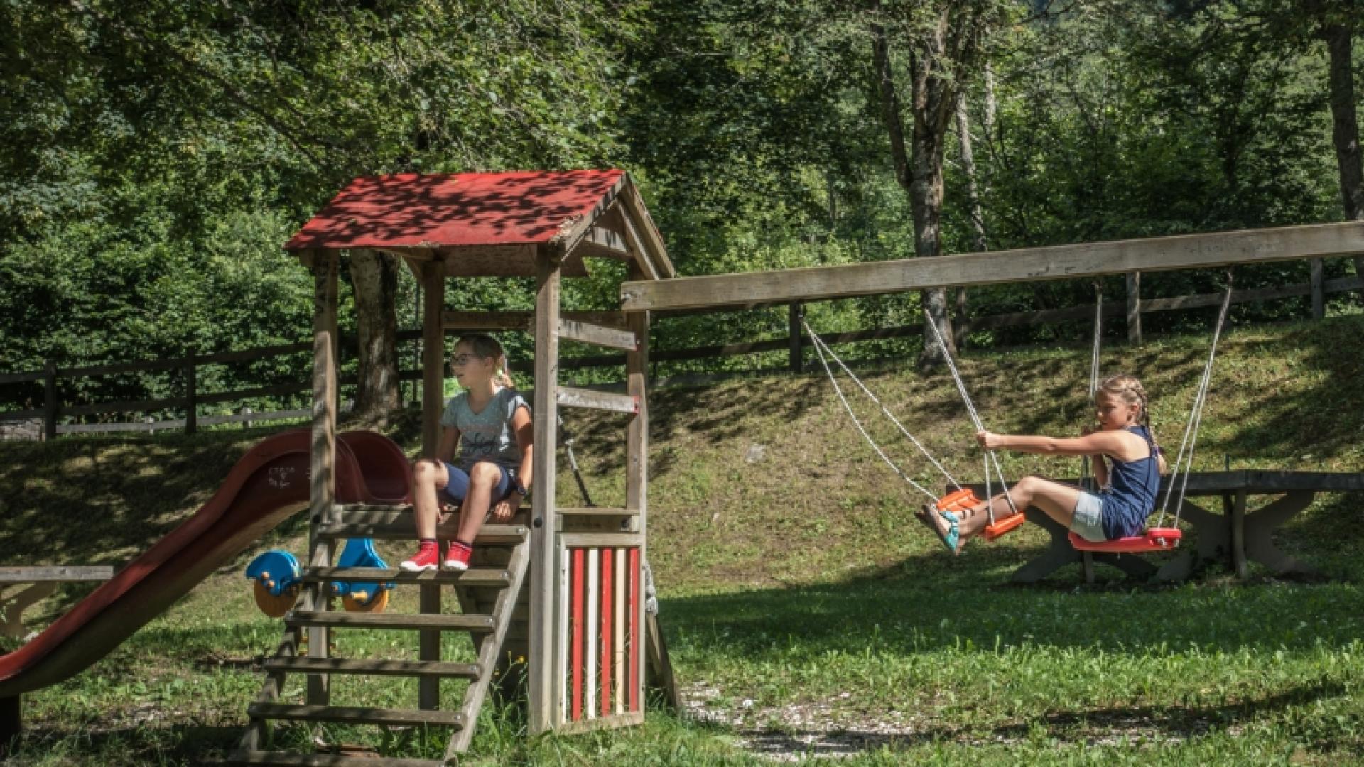 Bambini giocano su altalena e scivolo in un parco verde e soleggiato.